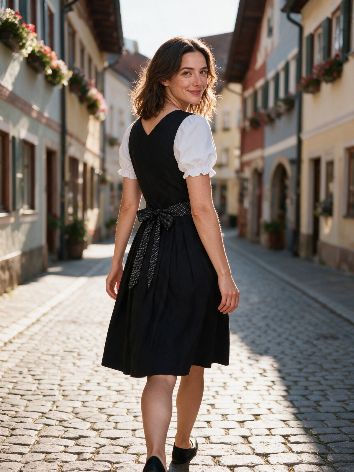 Woman walking an old-town street in the Lorelei black dirndl, polka-dot sash bow at the back
